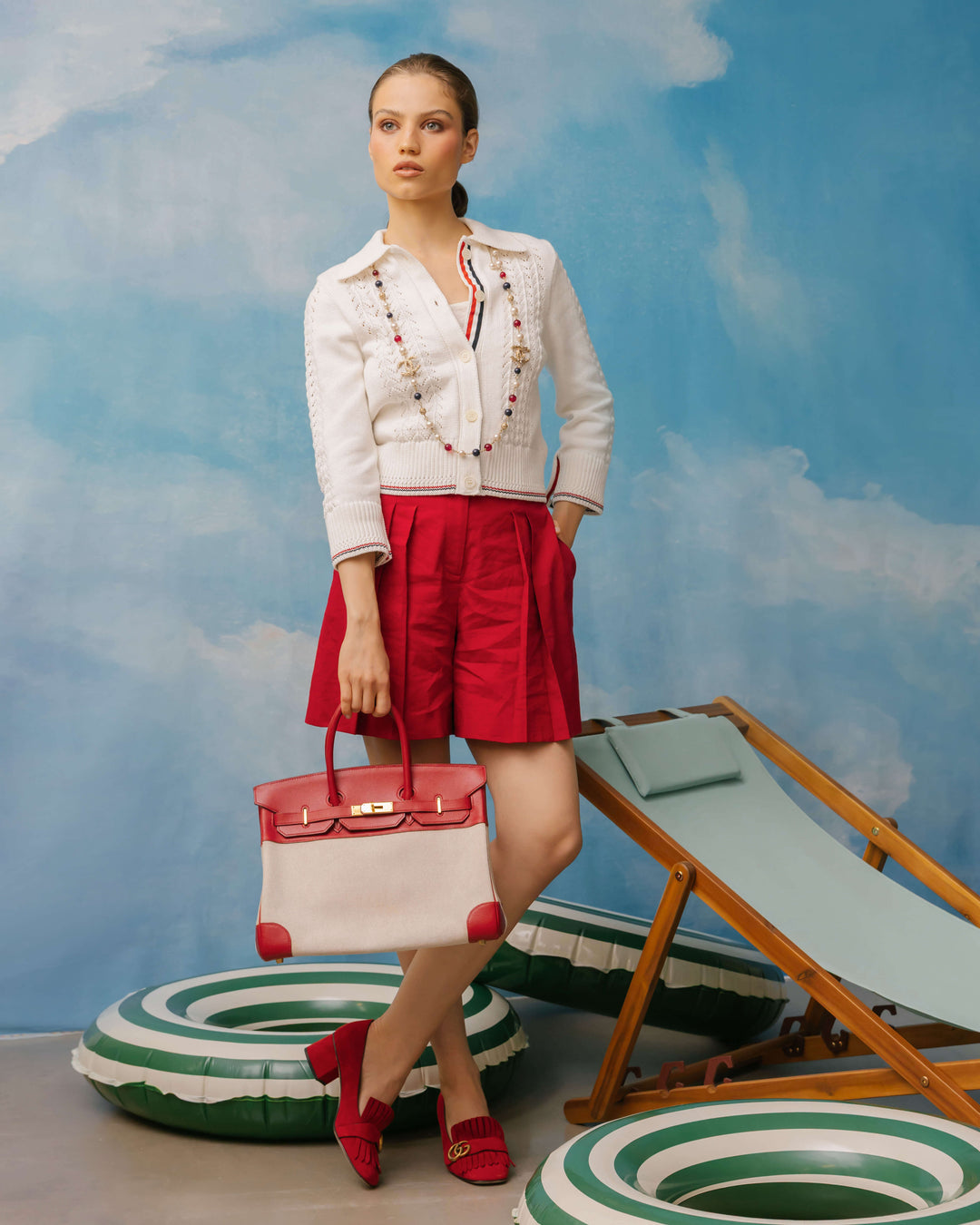 A woman stands in a studio setting wearing a white embroidered jacket, red shorts, and red shoes, holding a pink and white handbag. She is posed with green-and-white striped pool floats and a wooden lounge chair against a blue sky mural background.