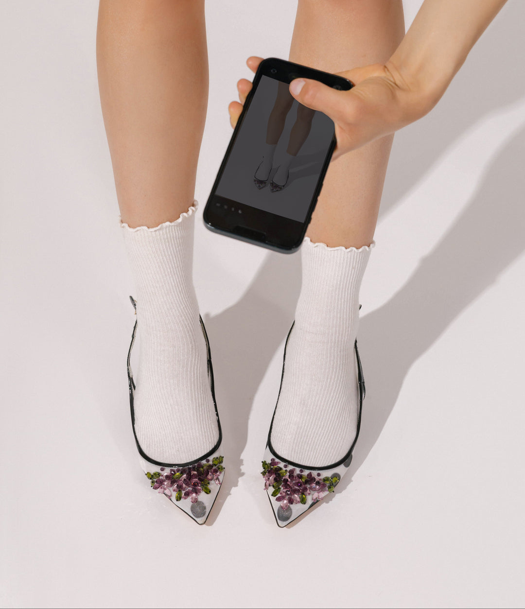A person's legs wearing white ribbed ankle socks and clear pointed-toe high heels with purple floral embellishments, while a hand holds a smartphone in front of the legs against a clean white studio background.