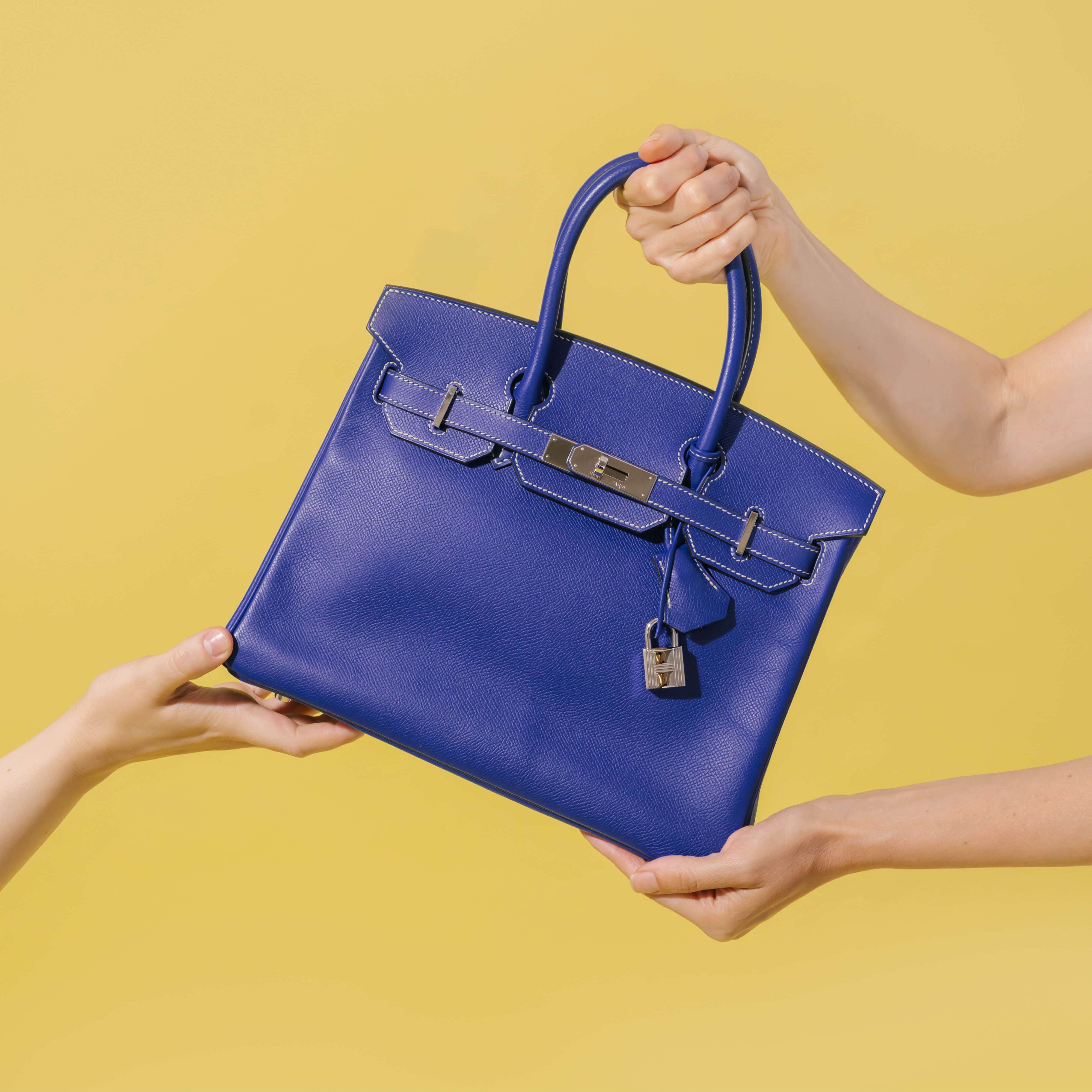 A royal blue leather handbag with gold-tone hardware is held by multiple hands against a solid yellow background.