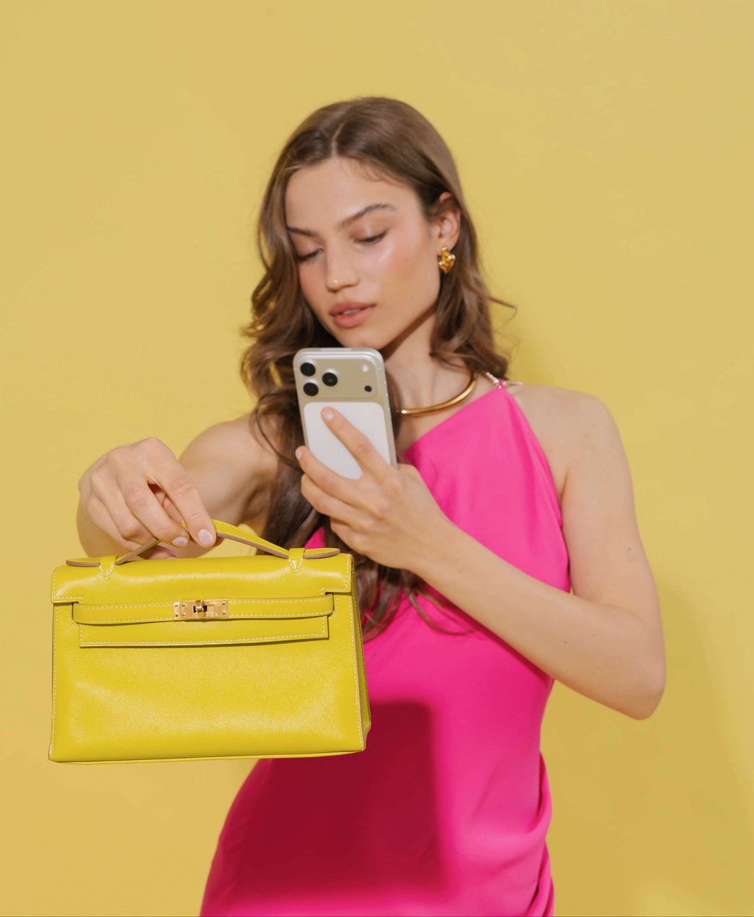 A woman in a bright pink dress holds a yellow handbag and a white smartphone against a solid yellow background.