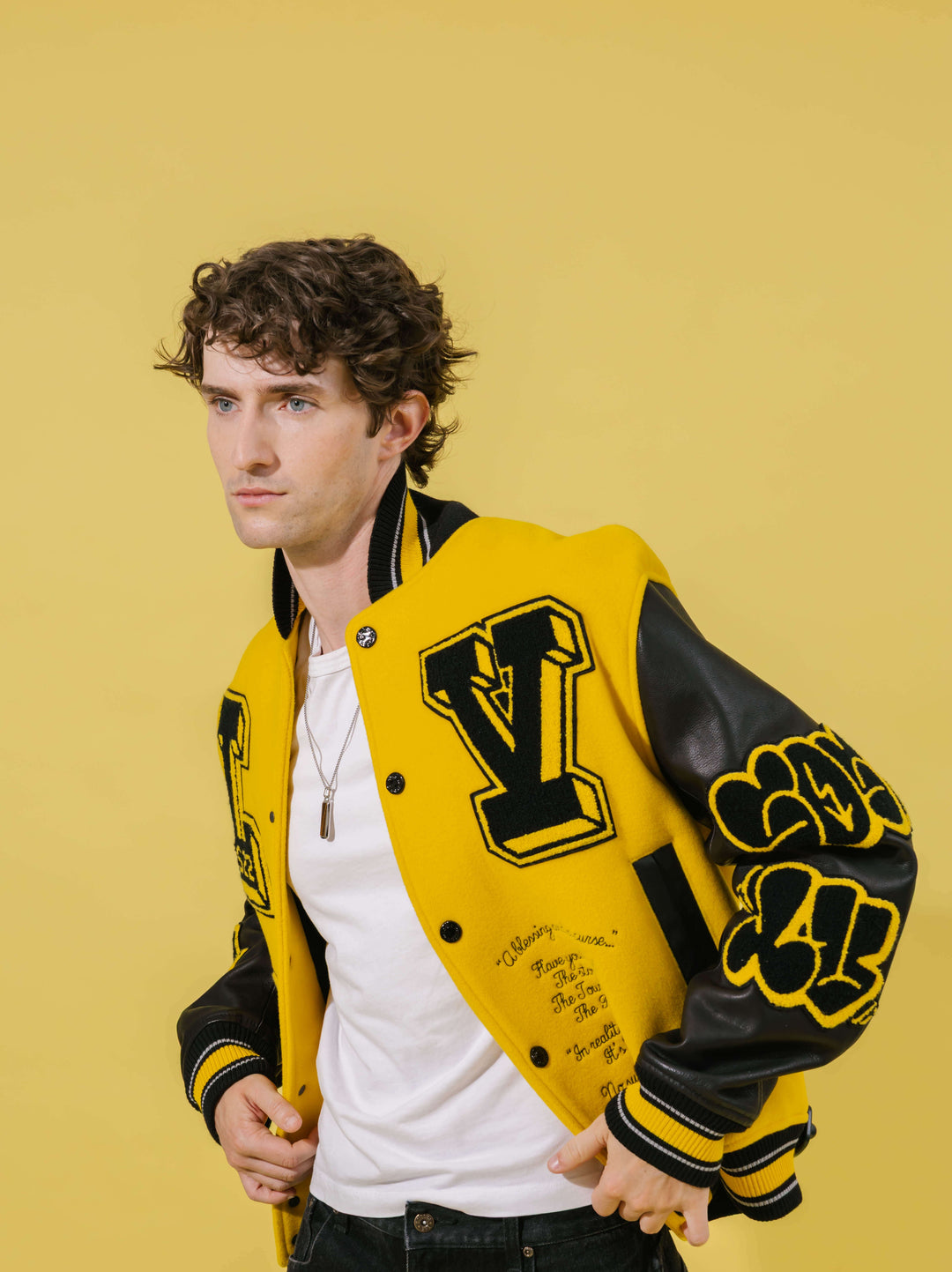 A man wearing a yellow and black varsity jacket with a large 'V' on the chest, posed against a solid yellow background.