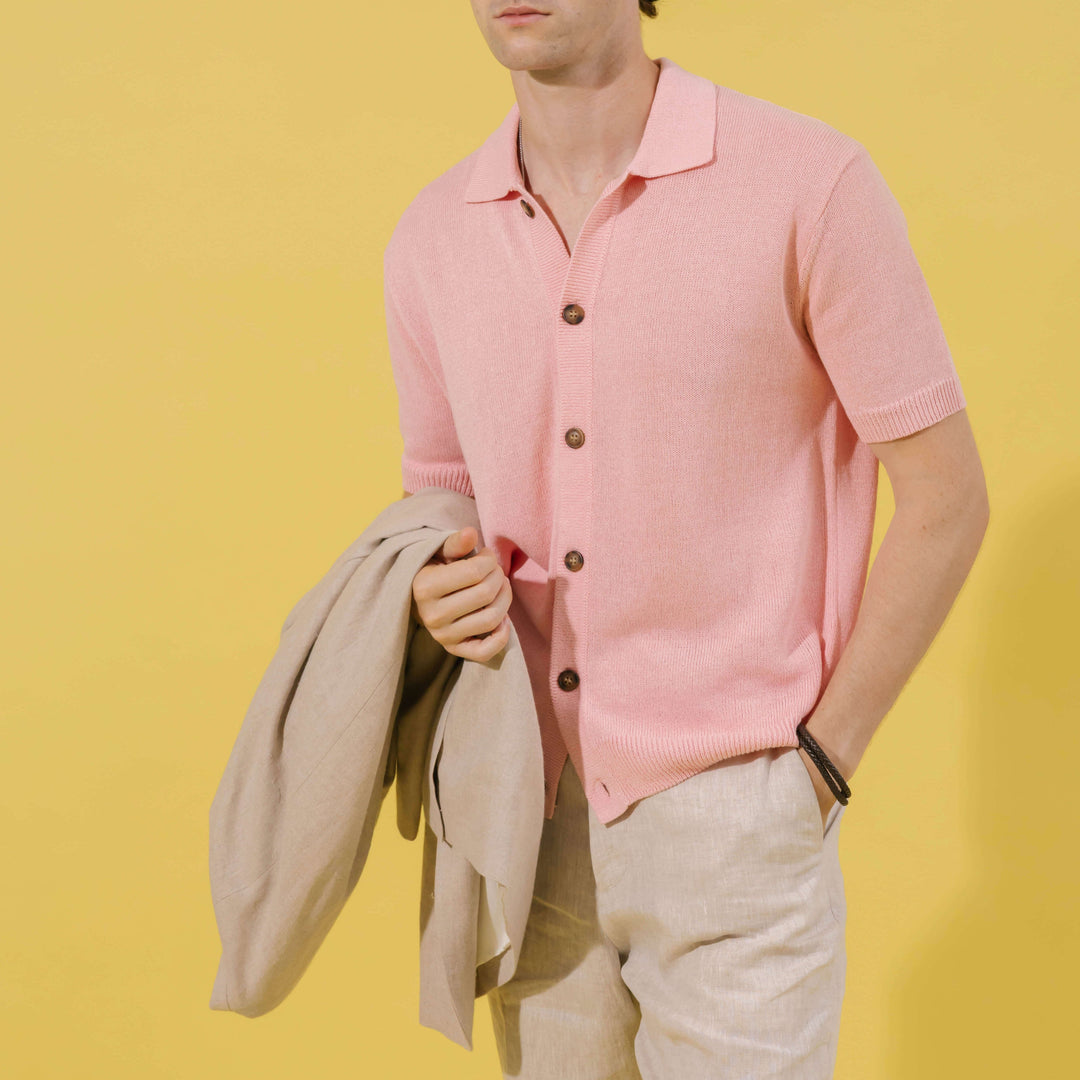 A man wearing a light pink short-sleeve knit polo shirt and beige trousers, holding a beige jacket against a solid yellow background.