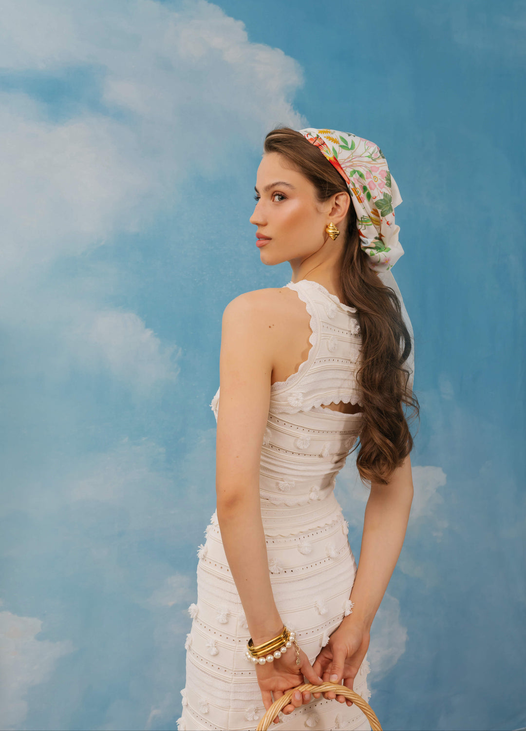 A woman in a white textured halter dress and a floral headscarf stands in profile against a blue sky backdrop with clouds. She is holding a woven basket and wearing gold jewelry.